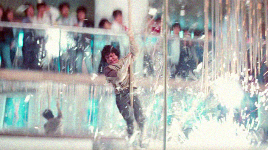 A man swings from a rope through shattering glass and bright sparks in a mall, with blurred onlookers and mirrored walls in the background, creating a dramatic action scene.