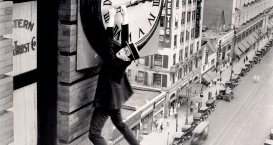 A man in a suit and hat hangs precariously from the hands of a large clock on the side of a tall building, above a busy city street filled with early 20th-century cars and pedestrians.