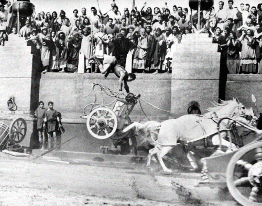 A dramatic moment in a chariot race; a man is thrown from his chariot as it crashes, while horses and other racers speed past, and a crowd of spectators watches from stone stands above.