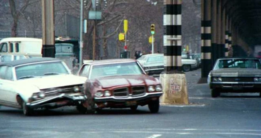 A white car and a red car collide on a city street under an elevated railway. Other vehicles are parked nearby, while leafless trees and traffic lights are visible in the background.