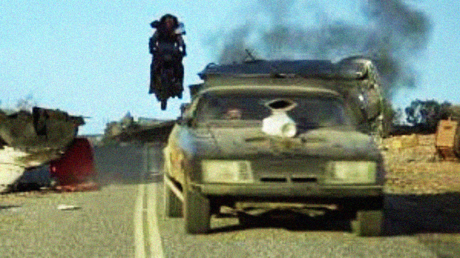 A person on a motorcycle jumps over a dusty, damaged car on a deserted road, with debris and smoke in the background under a clear blue sky.