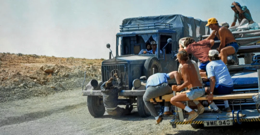 A film crew rides on the back of a truck, shooting a scene with another actor driving a blue truck on a dusty road. Crew members hold onto filming equipment while one person lies on the ground for a close-up shot.