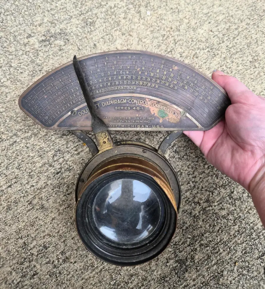 A hand holds an antique camera lens with a large glass element and a curved metal plate marked with detailed engraving and numbers, placed on a textured surface.
