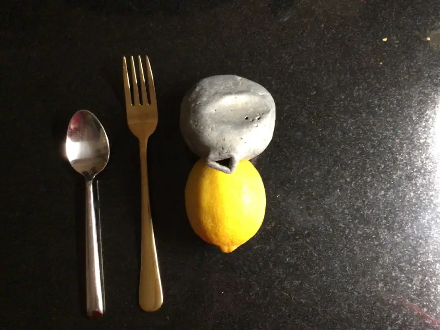 A spoon and a fork are placed side by side on a dark countertop. Next to them are a gray stone shaped like a lemon stacked on top of a real yellow lemon.