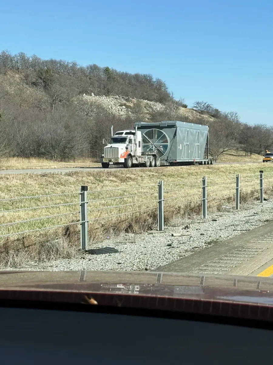 A white semi-truck with a large, rectangular metal structure on its trailer drives on a rural road, bordered by dry grass, a metal guardrail, and leafless trees under a clear blue sky.