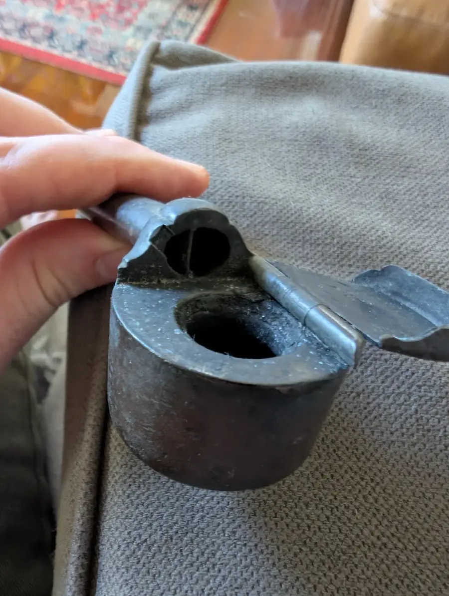 A hand holding a metal object with a cylindrical base and a hinged lid, featuring a circular hole in the center and another hole at the top. The object is resting on a textured gray fabric.