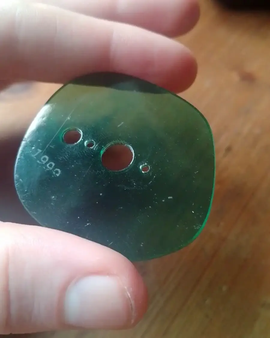 A hand holding a translucent, greenish plastic piece with four holes of varying sizes and the number "1993" printed on it, against a wooden surface background.