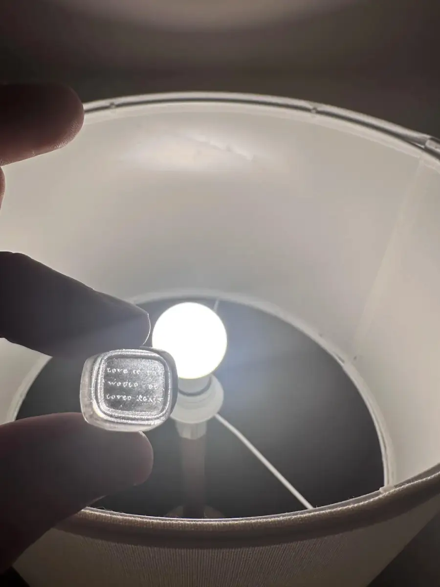 A hand holds a small item with engraved text near a lit light bulb inside a lampshade, the bright light illuminating the white lamp interior. The text on the item is difficult to read.