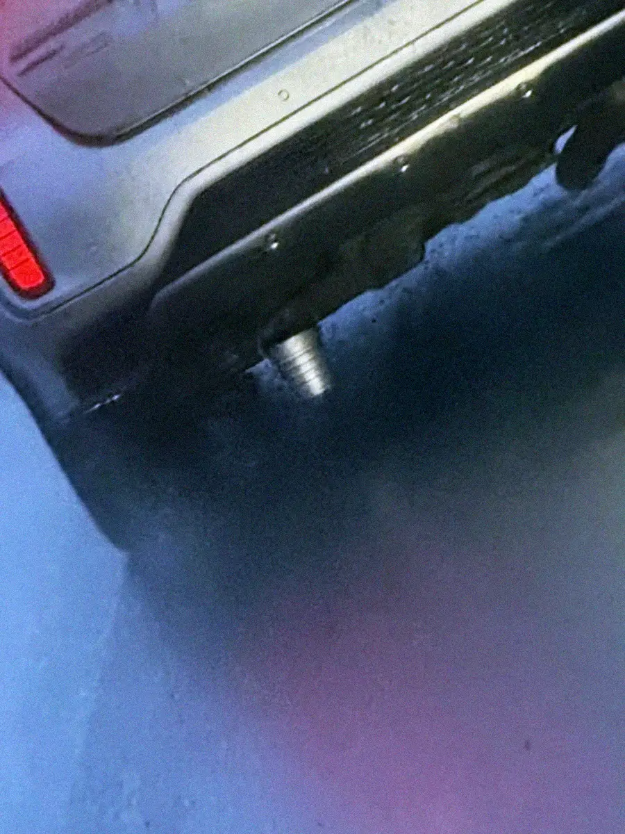 Close-up of the rear end of a car with a metallic spike attached underneath the bumper, parked on a wet surface. The left brake light is illuminated, and the car’s surface reflects the wet ground.