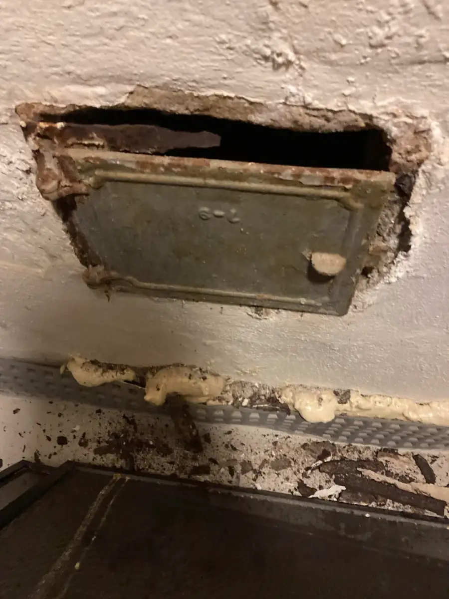 A rusty metal panel embedded in a chipped, white-painted wall with a rough opening around it; dirty pipes and debris are visible below the panel.