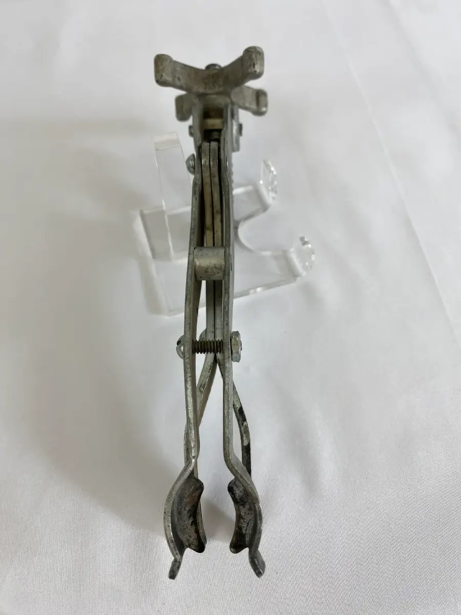 A vintage metal tool with two curved, tong-like ends and a spring mechanism, photographed standing upright on a white surface with a clear acrylic stand behind it.