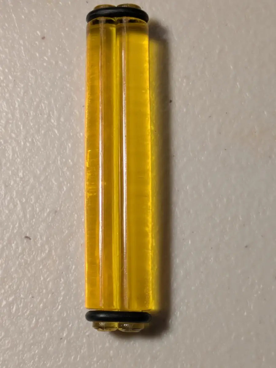 A close-up of a small, transparent yellow double-barreled plastic object with black rubber rings at both ends, placed vertically on a textured white surface.