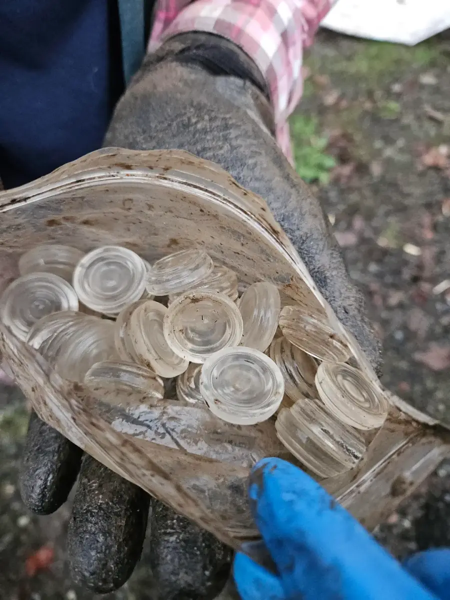 A gloved hand holds a dirty, transparent bag filled with several clear, circular glass objects. Another gloved hand points towards the bag. The background is an outdoor, earthy ground.