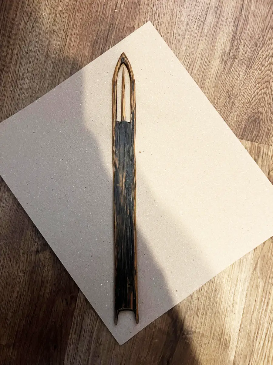 A dark, slender wooden object with a pointed top and two vertical cutouts lies on a square piece of light-colored paper, set on a wooden floor.