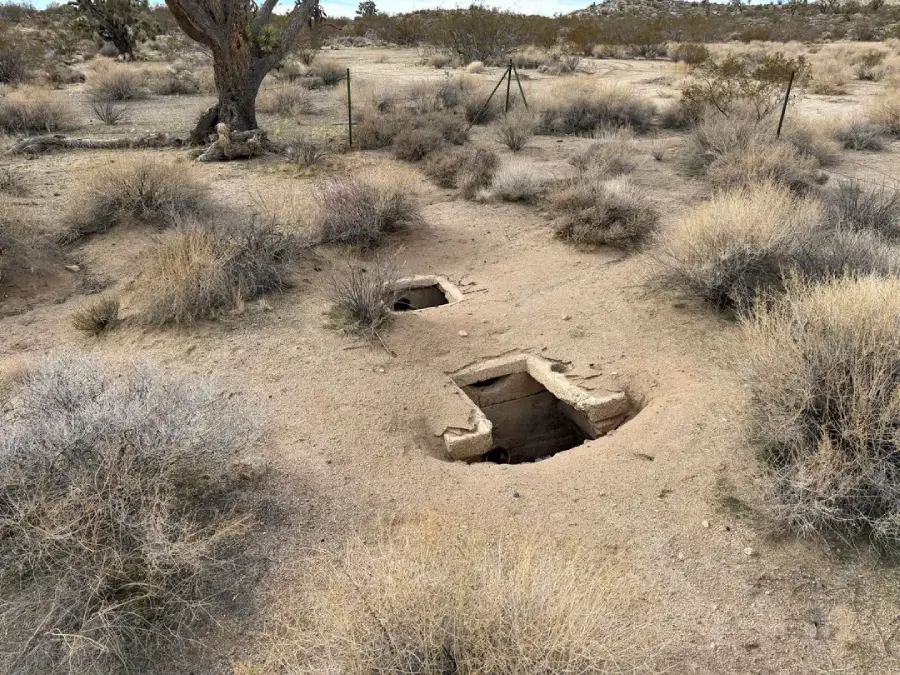 Two small, rectangular, concrete-lined openings in the ground in a dry, sparsely vegetated desert landscape, with shrubs and a tree nearby. A wire fence is visible in the background.