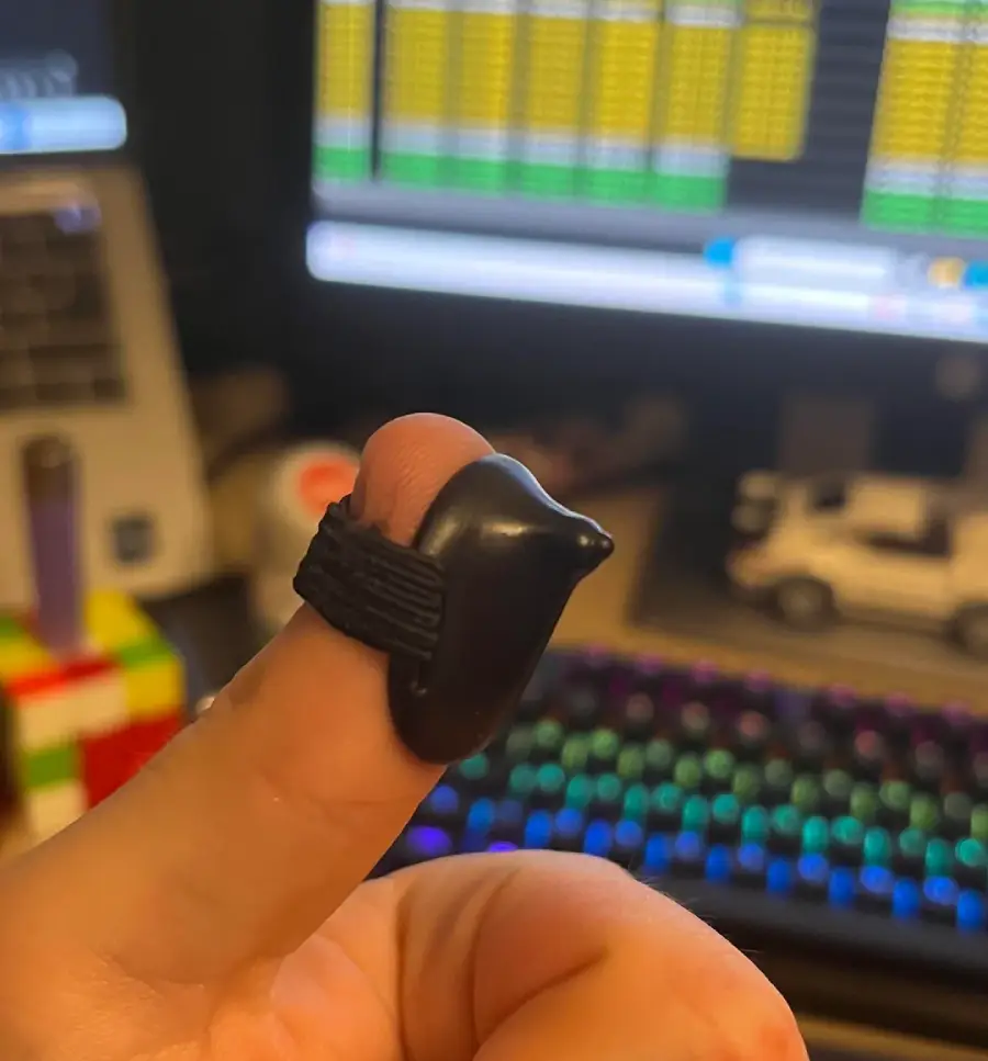 A thumb wearing a small black protective silicone cover, with a colorful keyboard, monitors displaying data, a toy car, and a Rubik's Cube in the blurred background.