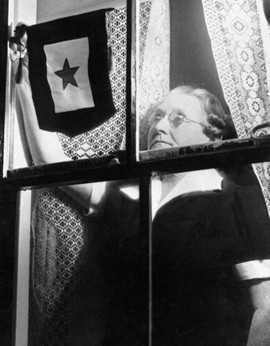 An elderly woman stands at a window, looking out while holding a “service flag” with a single star, indicating a family member serving in the military. Lace curtains frame the window in the black-and-white photo.