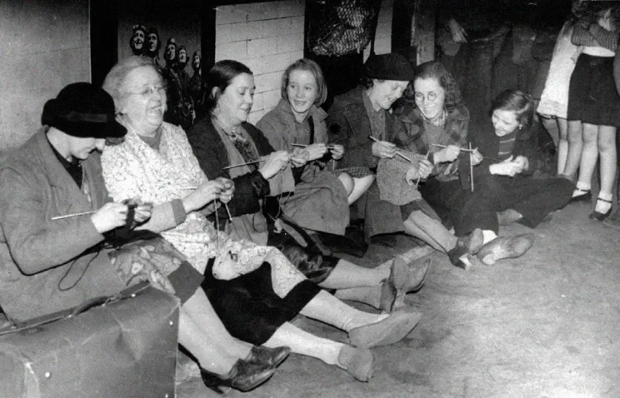 A group of women and girls sit in a row on the floor, knitting and smiling. Some older women appear to be teaching the younger ones. They are dressed in old-fashioned clothing, and their surroundings look modest and informal.