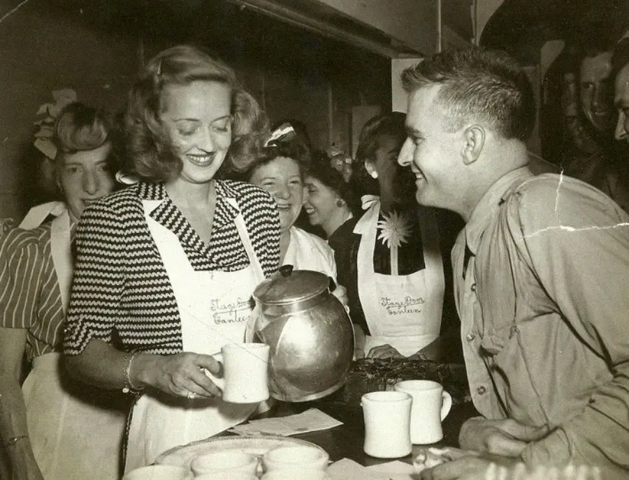 A smiling woman pours coffee into a mug for a uniformed man at a busy counter, surrounded by other cheerful people wearing aprons in a lively, vintage setting.