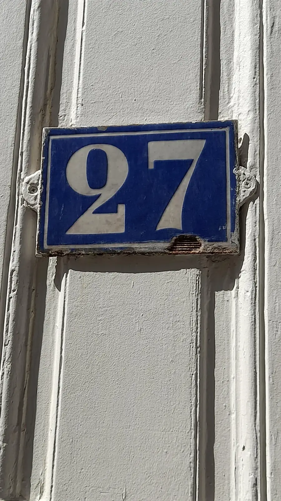 A rectangular blue and white enamel house number sign displays the number 27, attached to a light-colored, grooved wall with some visible chipping at the bottom edge.
