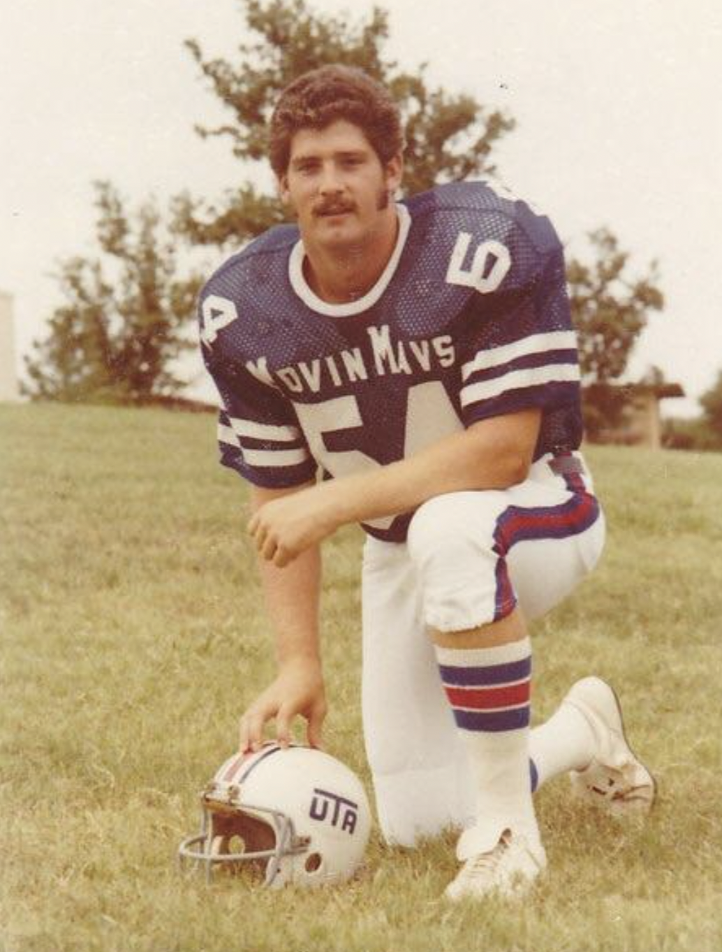 A football player in a blue jersey with “MAVS” and number 54 kneels on grass, holding a helmet labeled "UTA." He has dark hair, a mustache, and is outdoors with trees in the background.