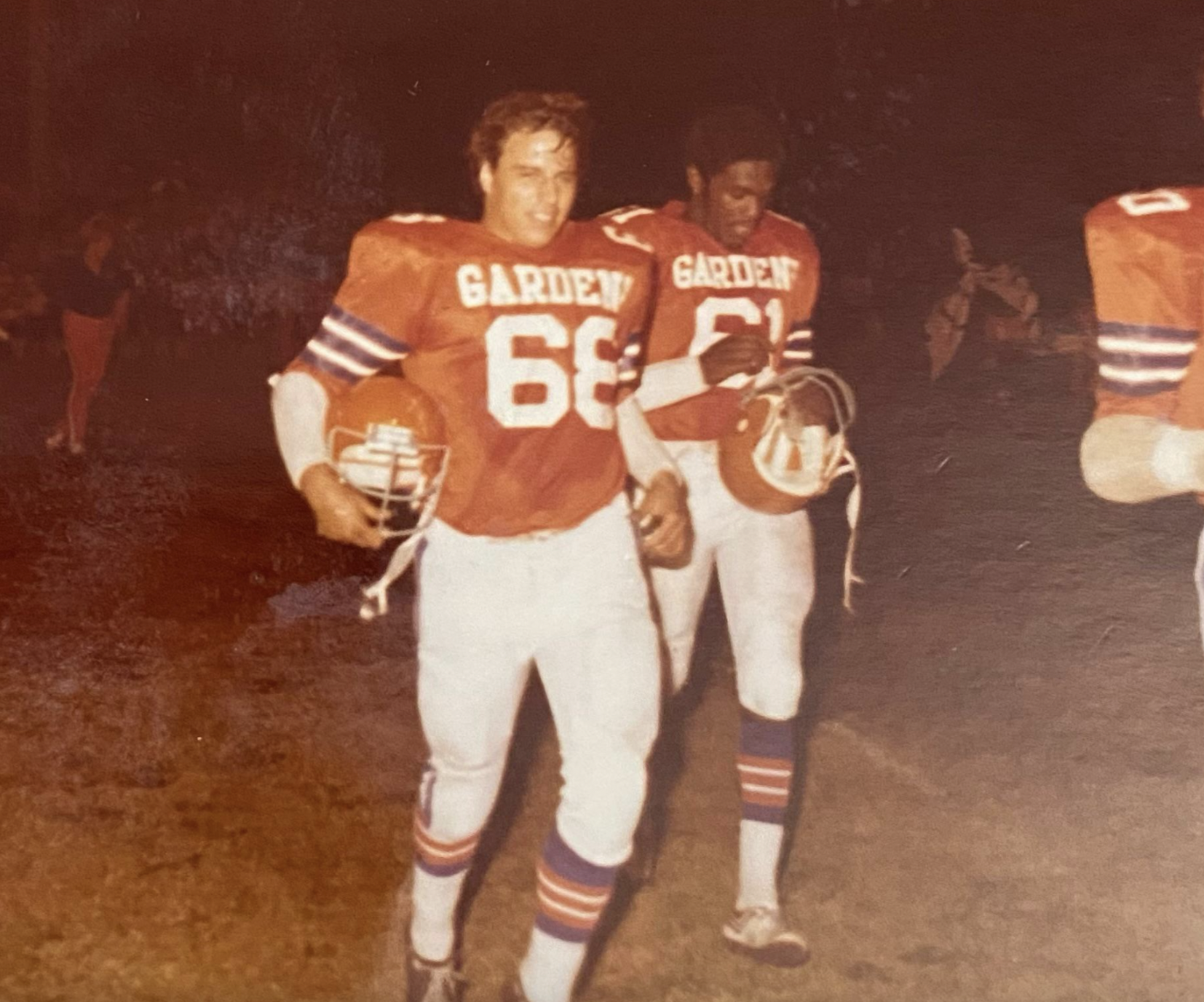 Two football players in orange "GARDEN" jerseys, numbers 66 and 61, walk off a grassy field at night, each holding a helmet and smiling. A few people are visible in the dimly lit background.