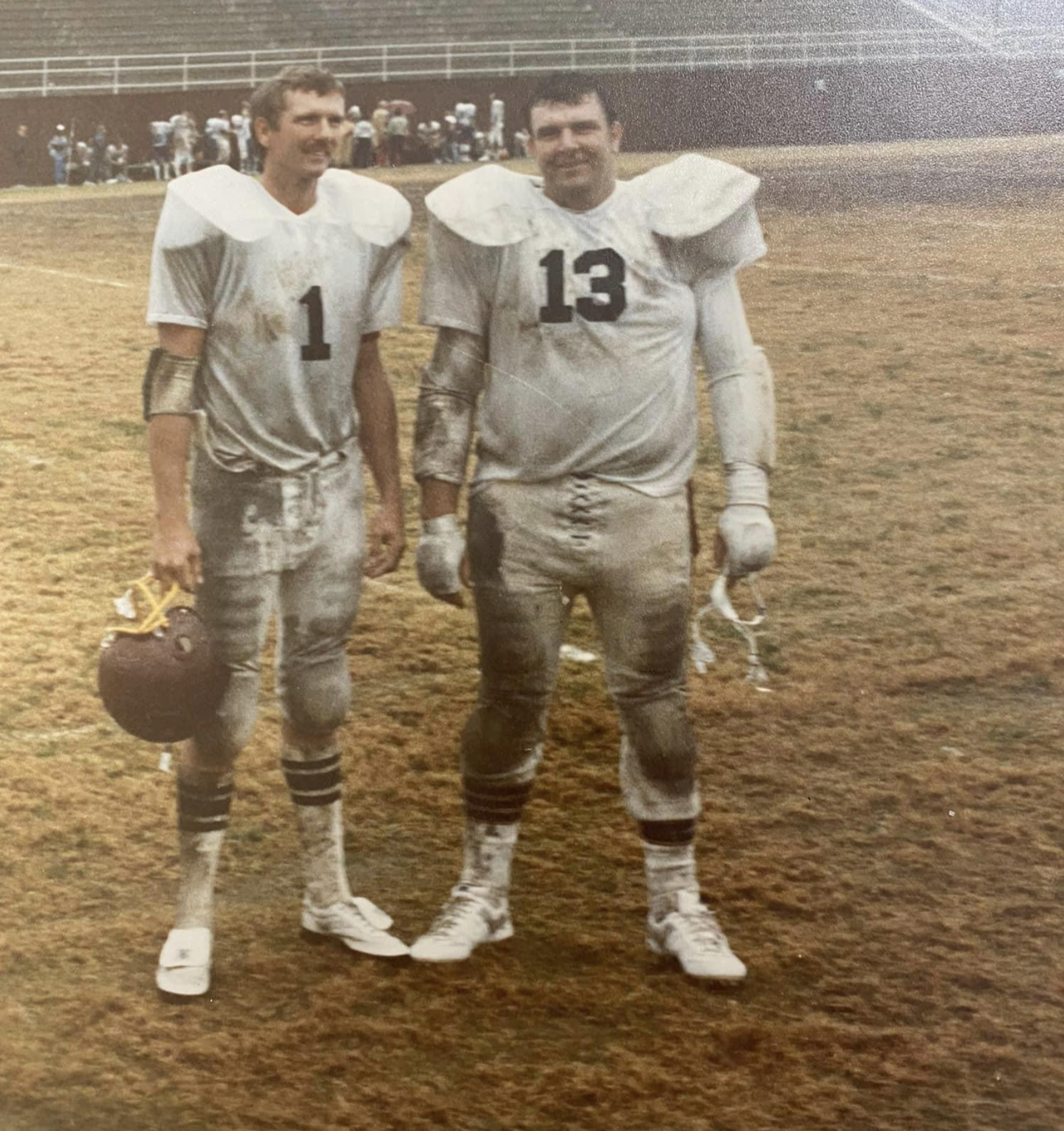 Two football players in white, muddy uniforms with numbers 1 and 13 stand on a worn football field, holding their helmets. Bleachers and other players are visible in the background.