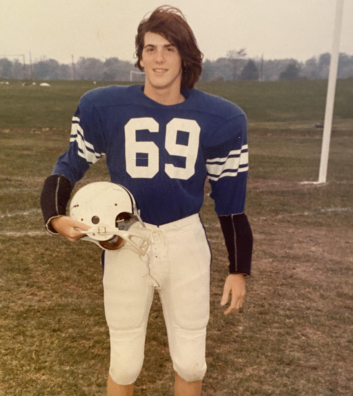 A young football player wearing a blue jersey with the number 69 and white pants stands on a grassy field, holding a white helmet and smiling at the camera.