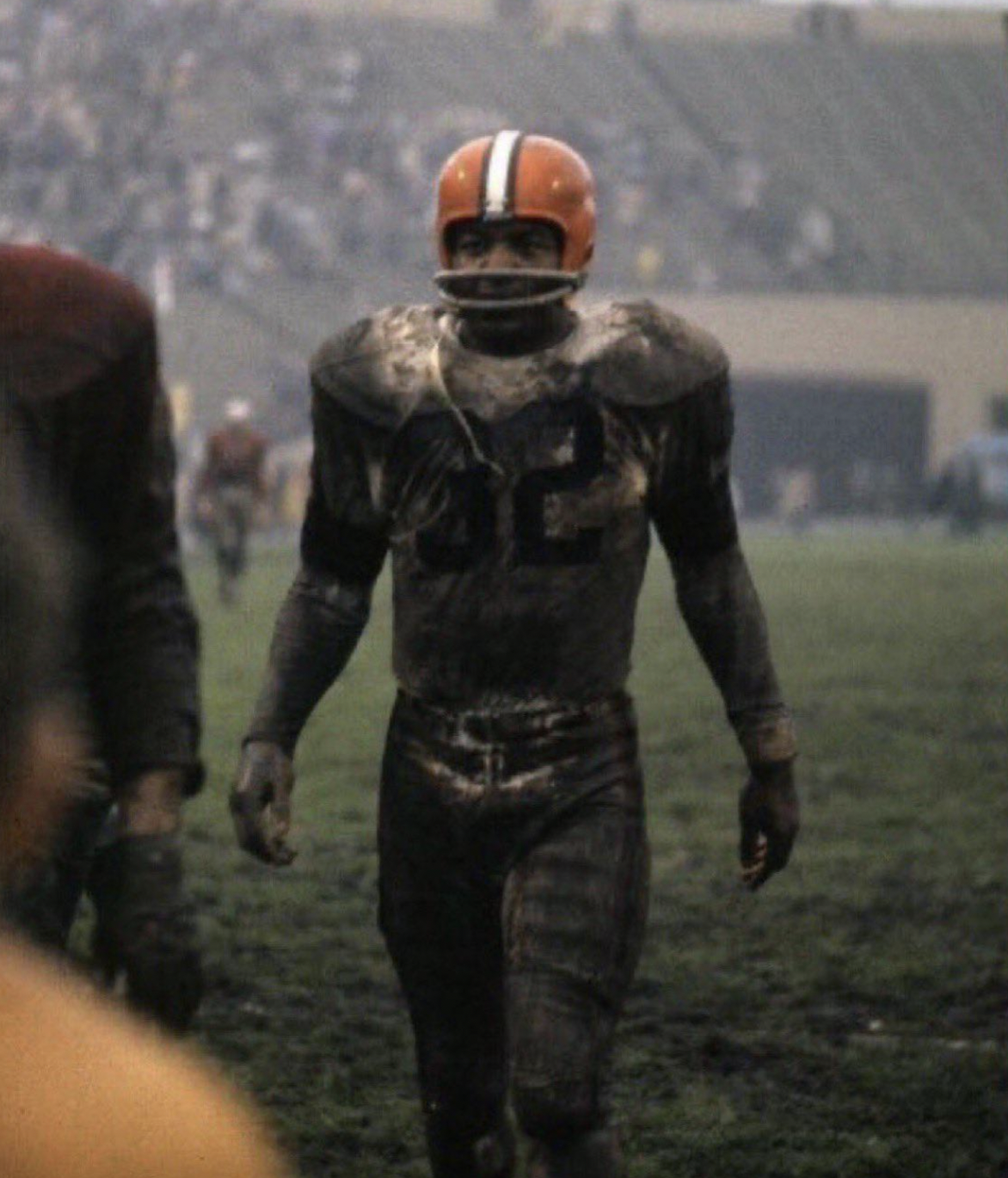 A football player in a muddy uniform and orange helmet walks on a grassy, dirt-covered field, with a stadium and other players visible in the background.