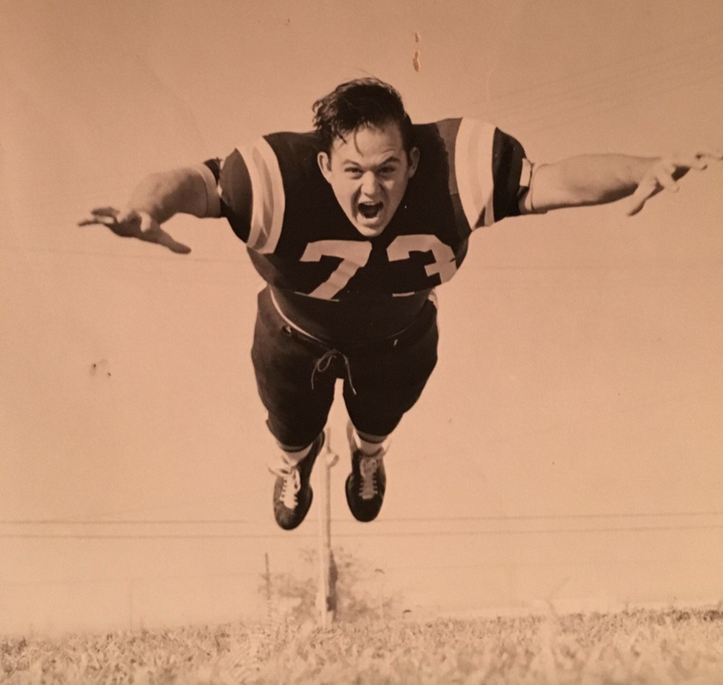 A vintage photo shows a football player in mid-air, wearing jersey number 73, arms outstretched and mouth open, as if diving or tackling on a grassy field.