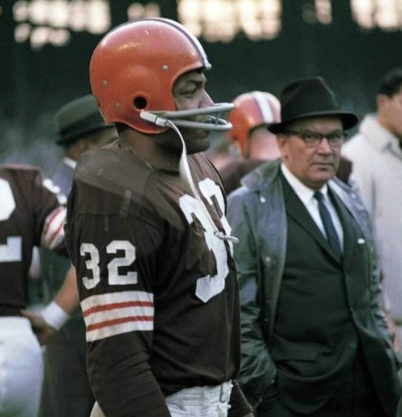 A football player wearing a brown uniform with number 32 and an orange helmet stands on the field, while a man in a suit, tie, glasses, and black hat stands nearby. Other people are visible in the background.