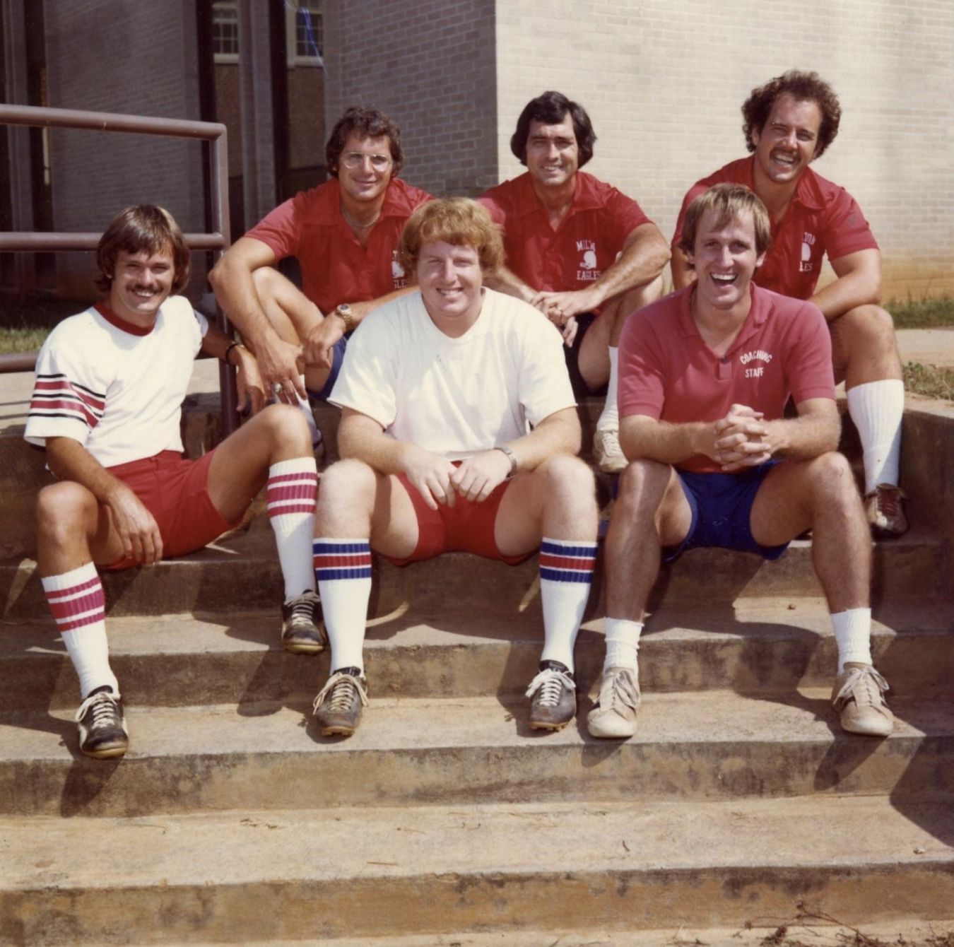 Six men sit on outdoor steps, smiling at the camera. Five wear matching red shirts and shorts; one in the center wears a white shirt and red shorts. All have athletic socks and sneakers. A building is visible in the background.