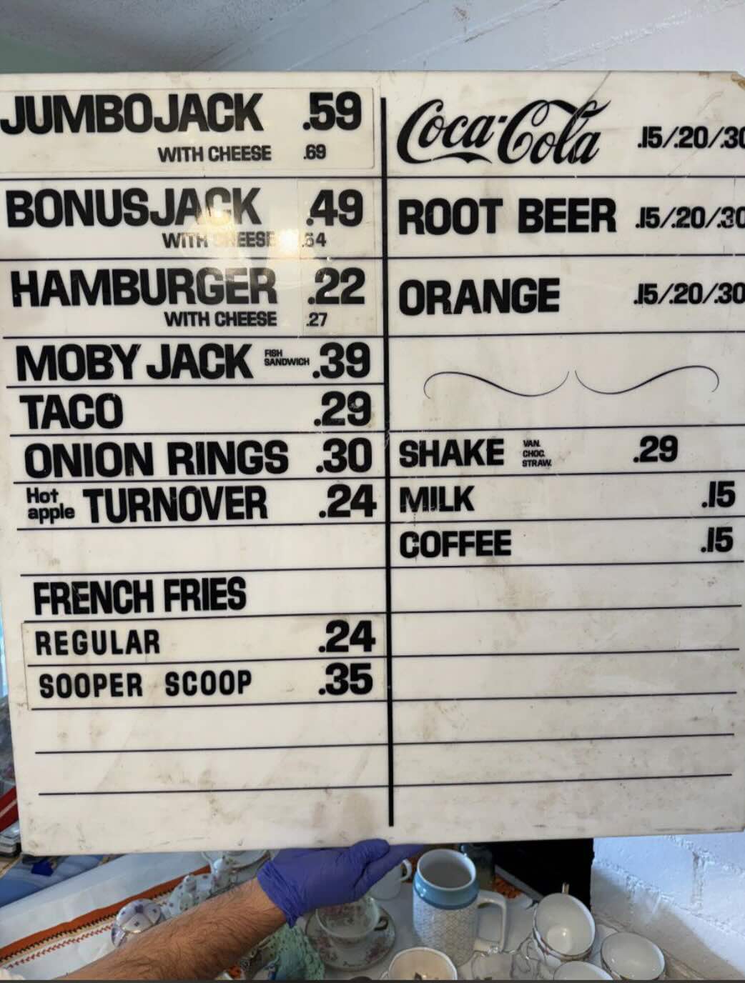 A vintage fast-food menu board displays items like Jumbo Jack, Hamburger, Moby Jack, Taco, onion rings, and drinks, along with their prices. A gloved hand holds the bottom corner of the board.
