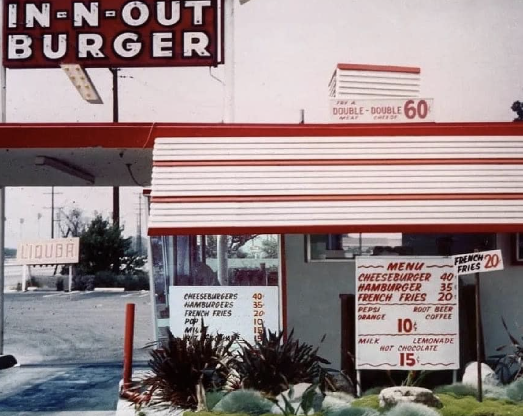 Vintage In-N-Out Burger stand with a large sign and striped awning, displaying a menu board listing cheeseburgers, hamburgers, fries, and drinks with prices in cents. Shrubs are in the foreground.
