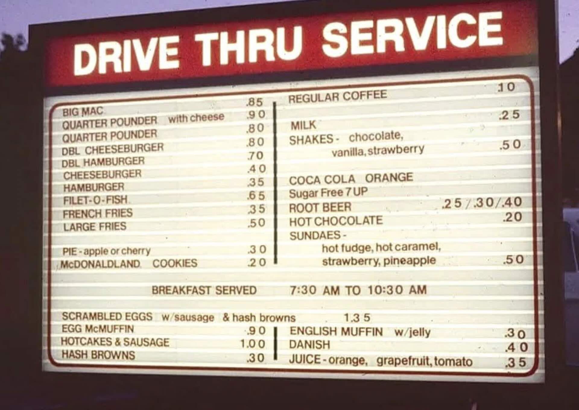 A drive-thru menu board displays various food and drink options, including burgers, fries, breakfast items, desserts, and beverages, with prices listed beside each item. The sign is illuminated, making the text visible.