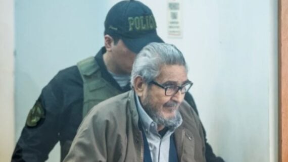 An elderly man with gray hair, glasses, and a beard is escorted by a uniformed police officer through a doorway. The elderly man appears to be smiling slightly.