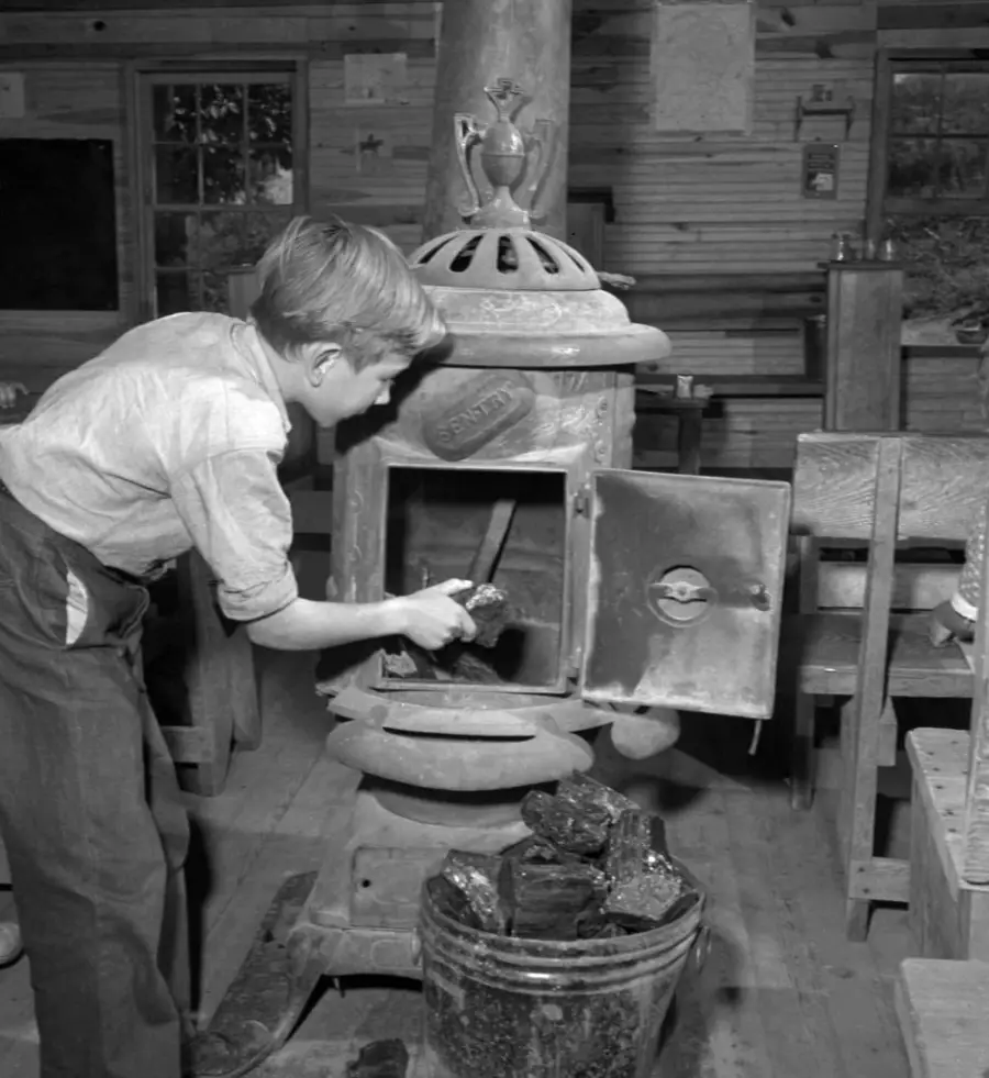 A boy in rolled-up sleeves adds coal to a potbelly stove inside a rustic wooden room. A metal bucket filled with coal sits nearby, and wooden benches and walls are visible in the background.