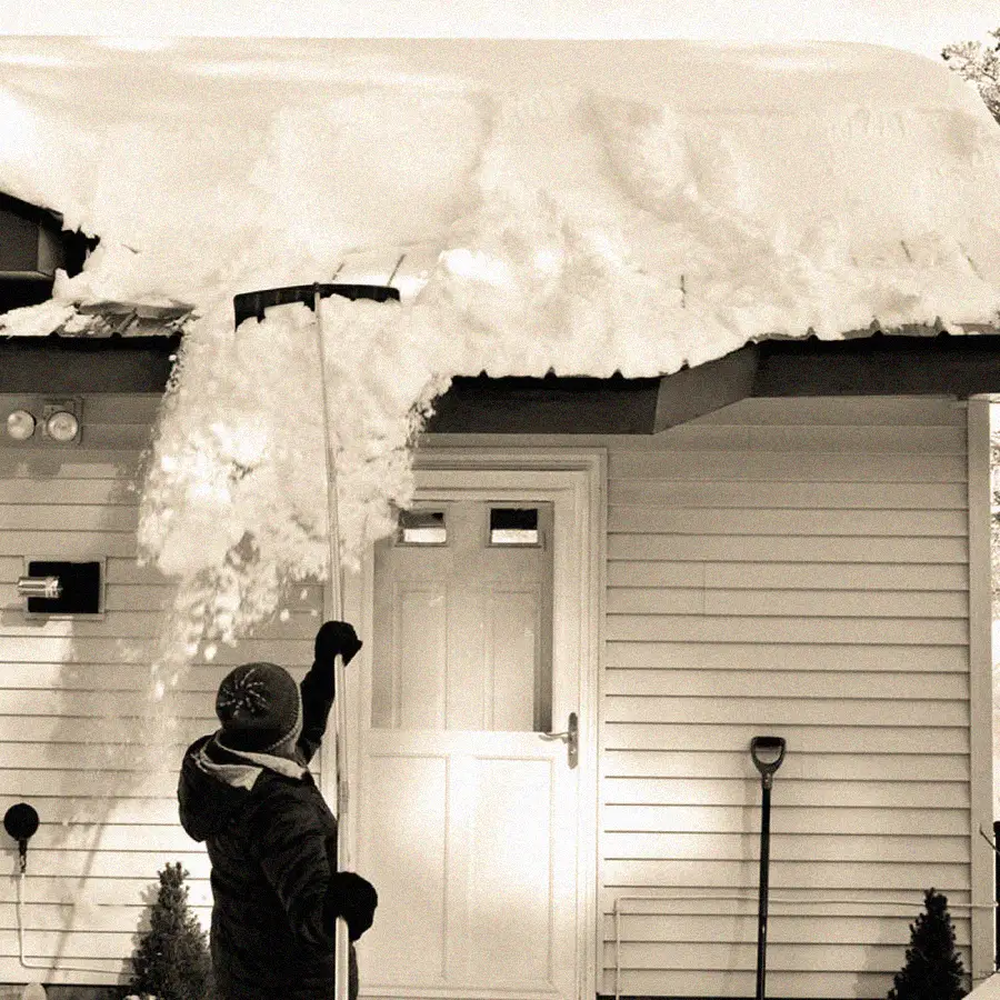 A person wearing a winter hat uses a long tool to remove thick snow from the roof of a house. Snow falls from the roof while the person stands near the front door. A snow shovel leans against the house.