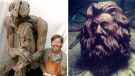 A smiling artist poses next to a large, unfinished clay sculpture of a seated, contemplative figure. Beside this, there is a detailed clay bust of a bearded man with long hair on a worktable.