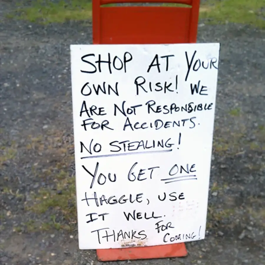 A handwritten sign on a white board reads: "Shop at your own risk! We are not responsible for accidents. No stealing! You get one haggle, use it well. Thanks for coming!" The sign is outdoors on gravel.