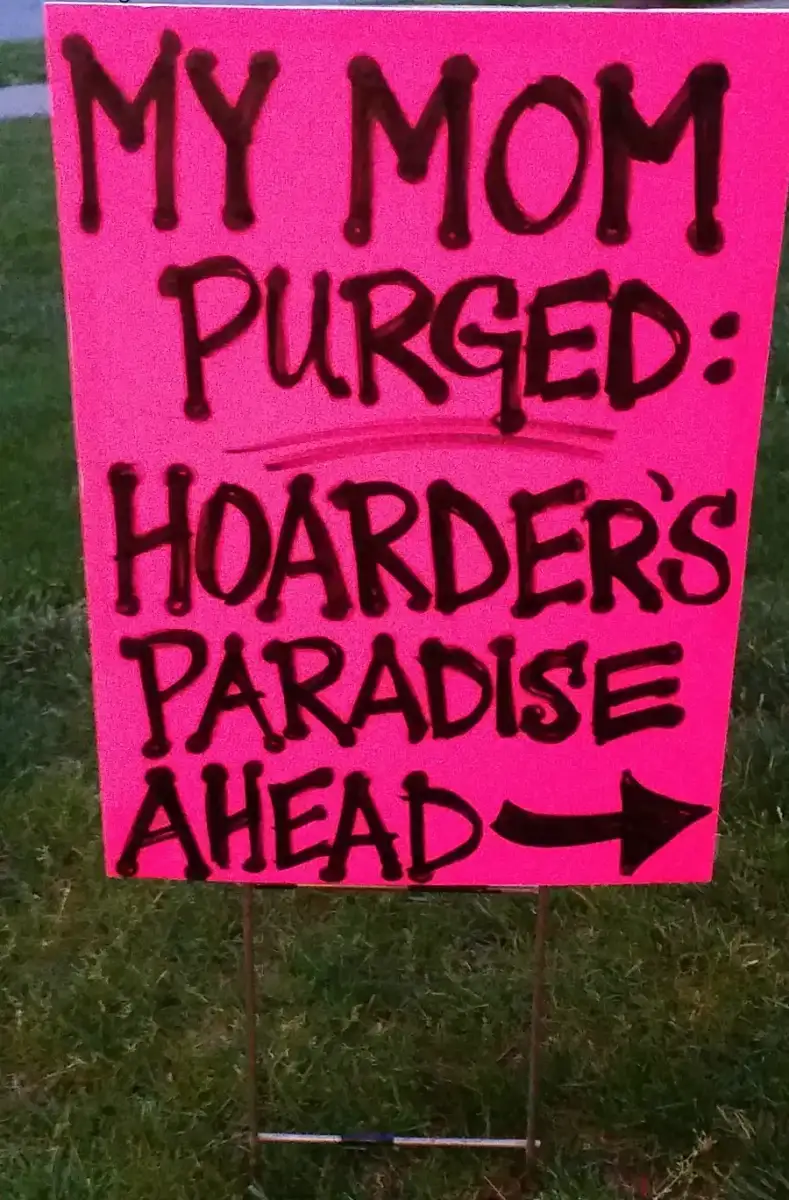 A bright pink yard sign with bold black lettering reads, "MY MOM PURGED: HOARDER'S PARADISE AHEAD" with a large arrow pointing to the right. The sign is placed on grass.
