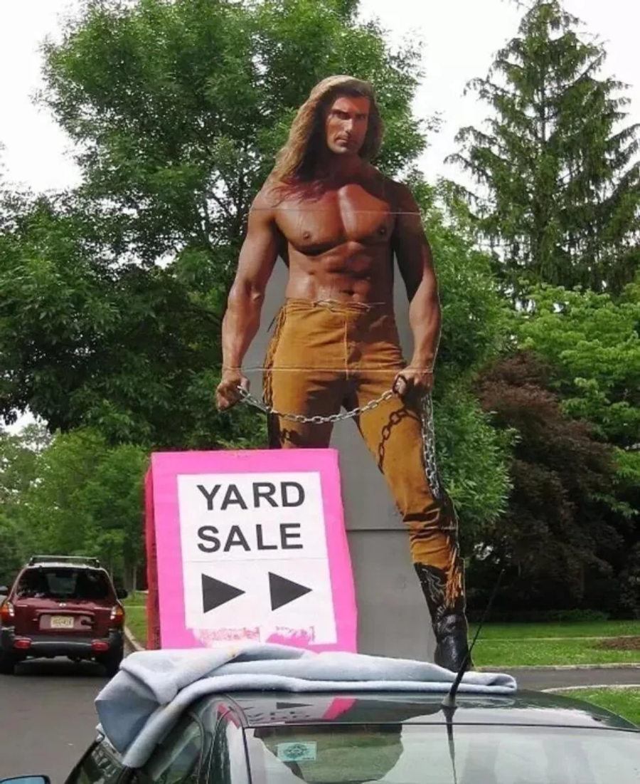 A cardboard cutout of a shirtless, muscular man with long hair stands behind a car. A pink "Yard Sale" sign with black arrows is attached to the car's windshield. Trees and parked vehicles are in the background.