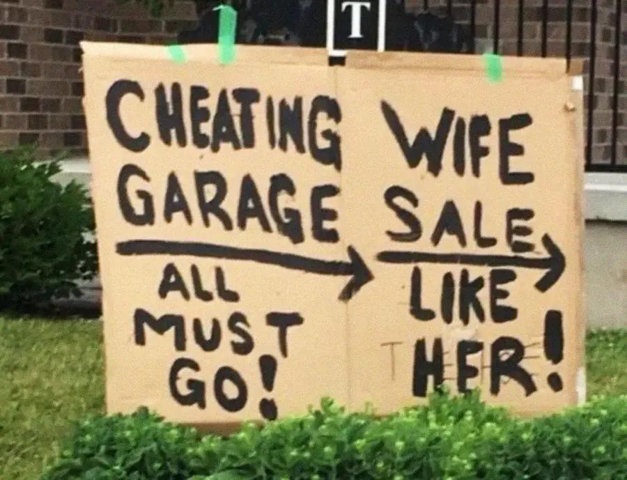 Two cardboard signs in a yard read: "CHEATING WIFE GARAGE SALE – ALL MUST GO! --> LIKE HER!" with bold black letters and arrows pointing right. Shrubs and a brick building are visible in the background.