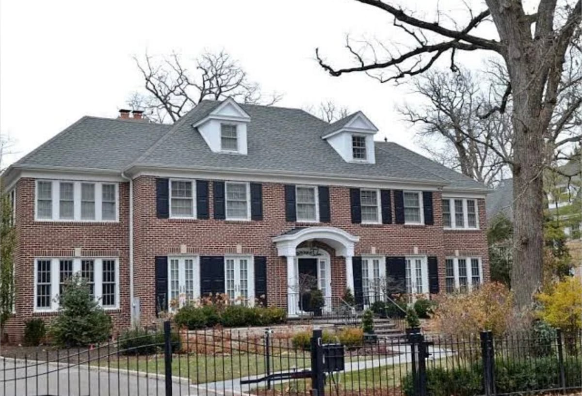 A large two-story brick house with black shutters, white trim, and a gray roof, featuring dormer windows, a decorative arched entryway, and a gated front yard with shrubs and bare trees.