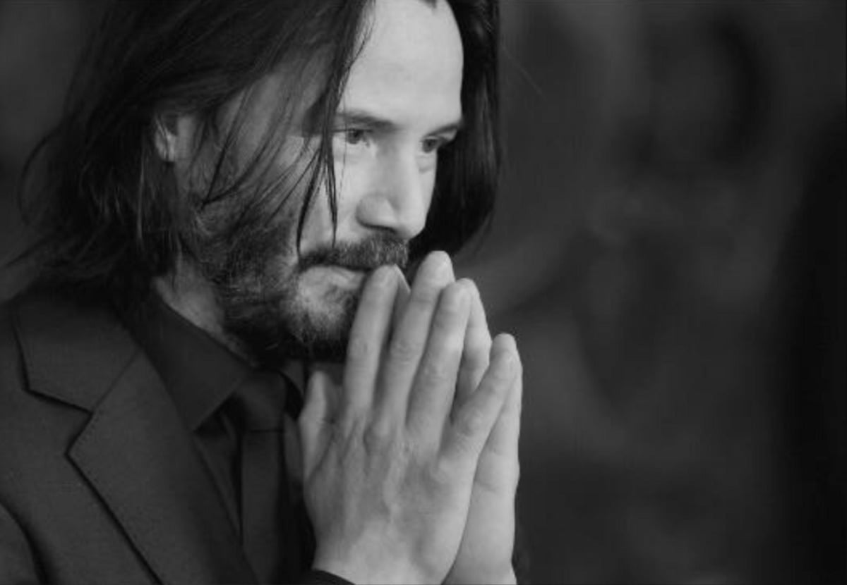 A man with shoulder-length dark hair and a beard, wearing a suit, clasps his hands together near his face in a thoughtful or contemplative pose. The image is in black and white.