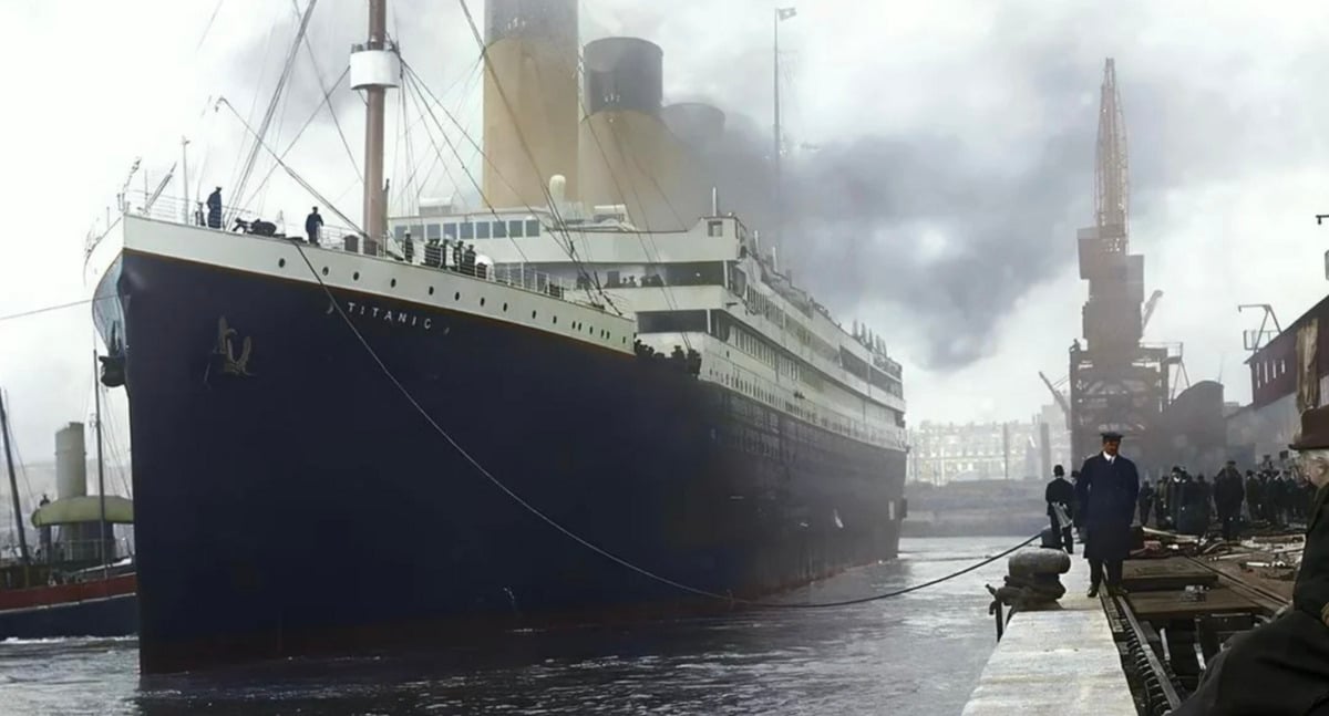 The RMS Titanic, a large ocean liner, docked at a port with smoke rising from its funnels. People and workers stand nearby, and industrial cranes are visible in the background.