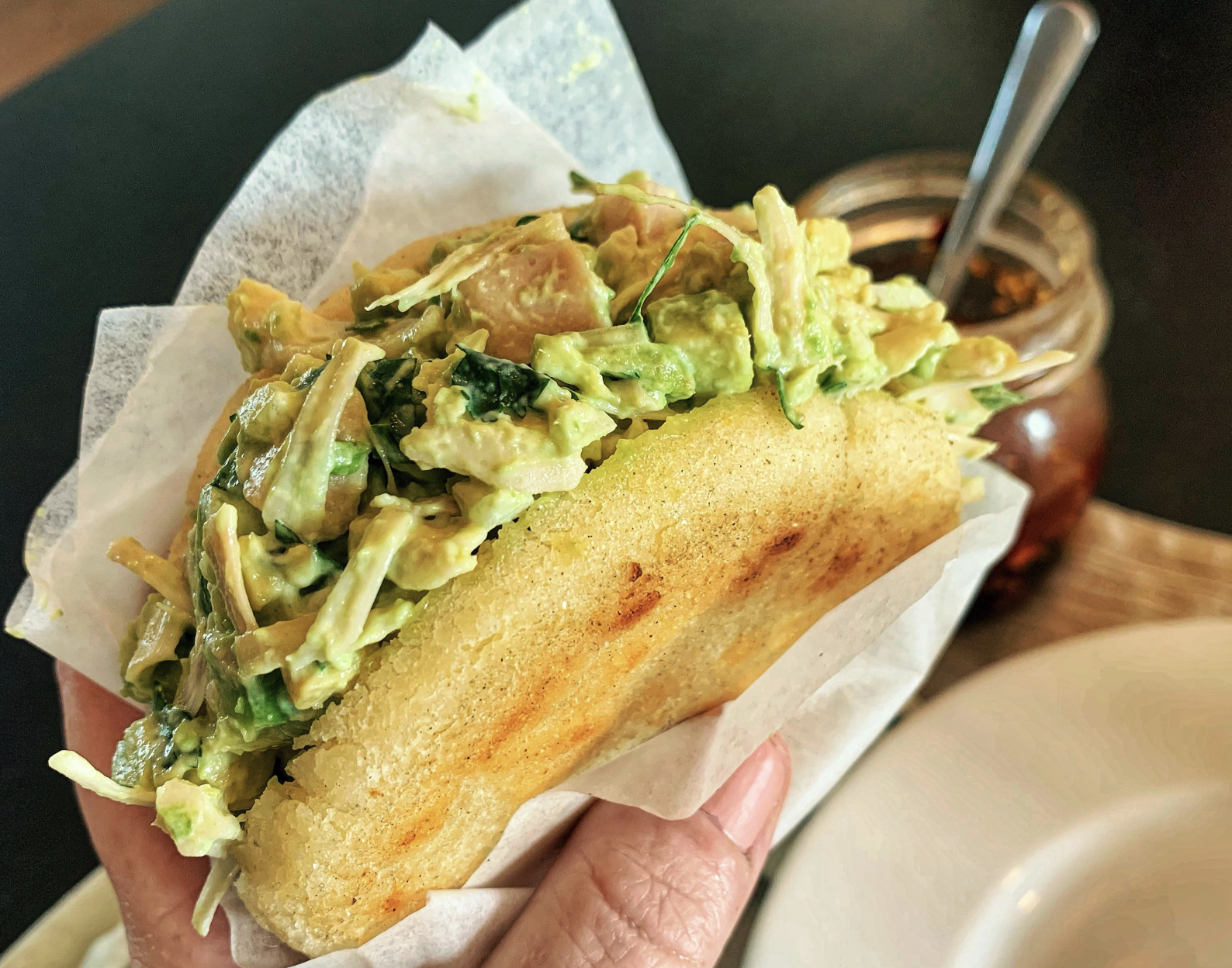 A hand holds an arepa stuffed with a creamy mixture of avocado, chicken, and herbs, wrapped in white paper. In the background, there is a glass jar with a spoon and part of a white plate.