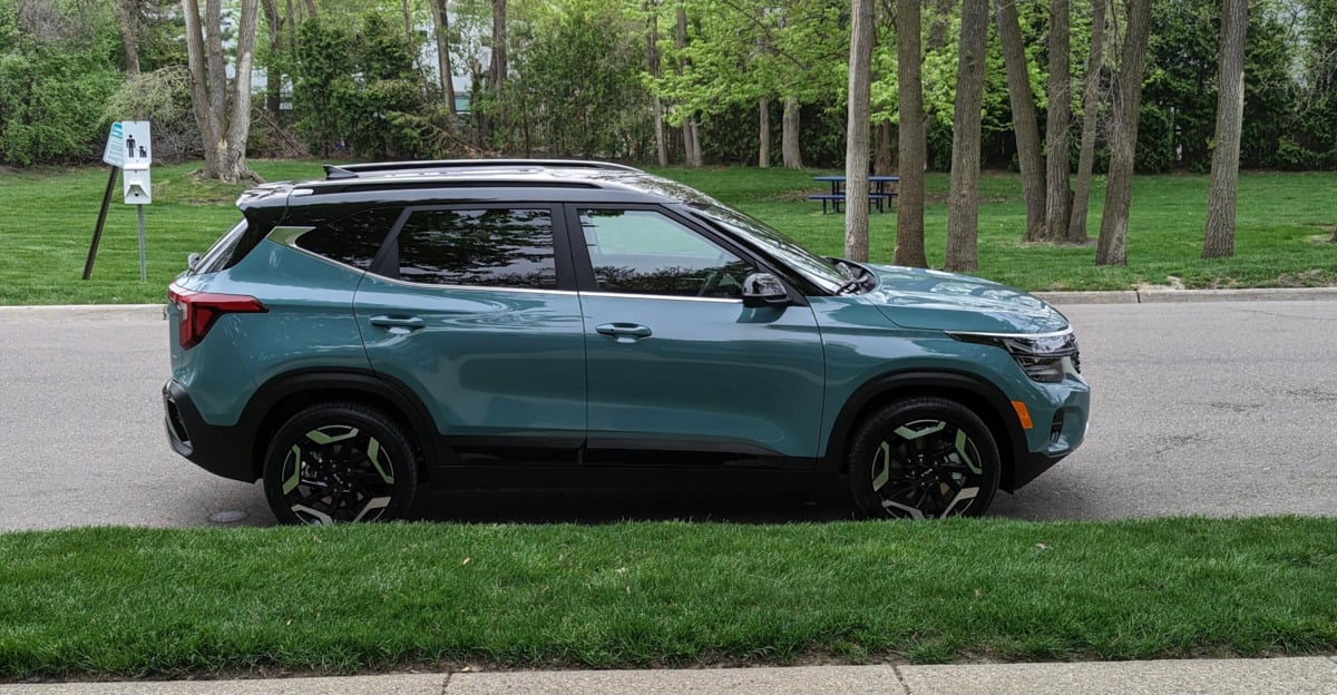 A teal SUV is parked on a street beside a grassy area, with trees and a picnic table in the background.