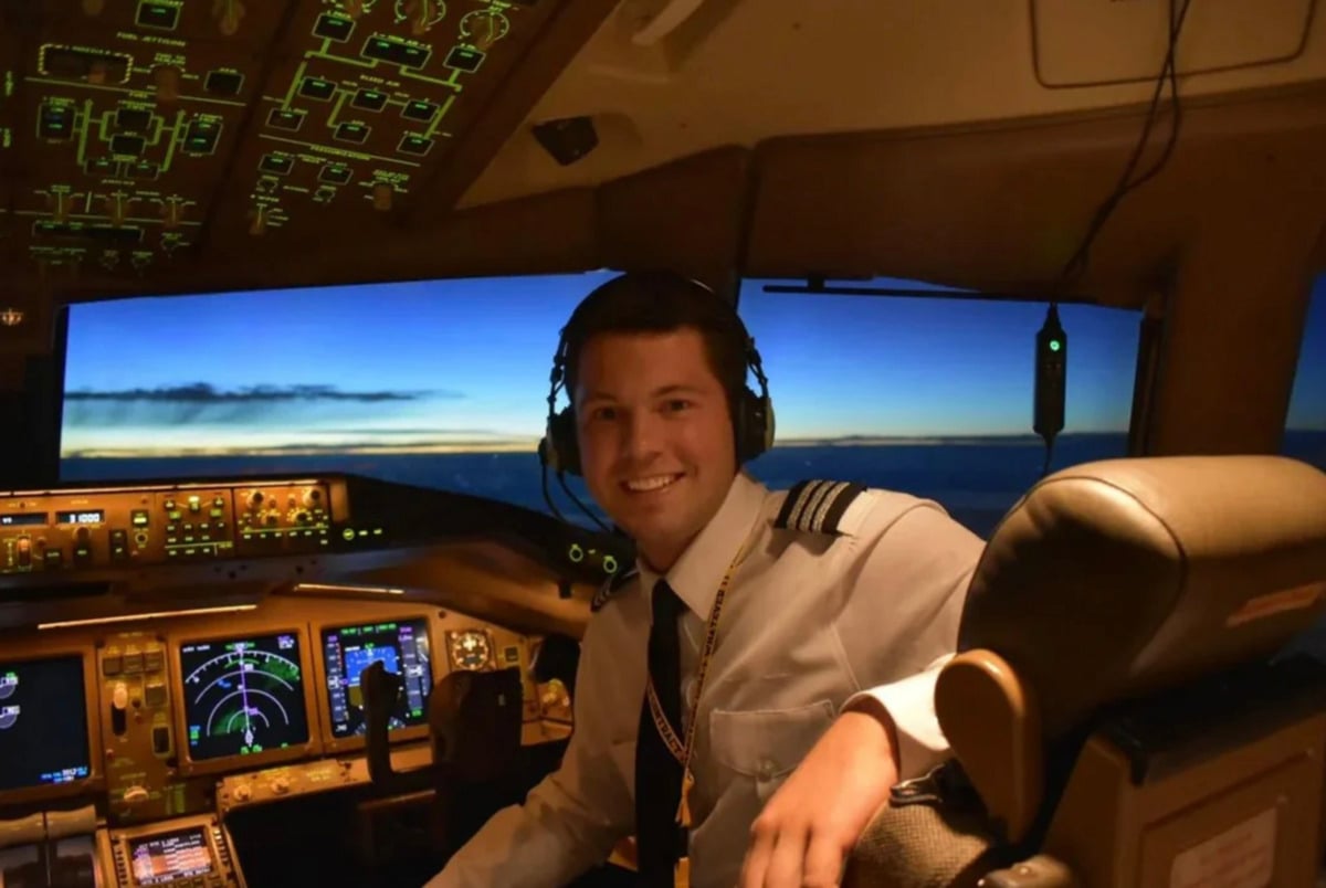 A smiling pilot wearing a headset and uniform sits in an airplane cockpit, surrounded by illuminated controls and screens, with a scenic view of the sky visible through the windows.