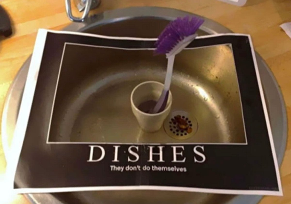 A kitchen sink with a dishwashing brush in a cup, and a paper sign draped over the sink that reads, "DISHES: They don't do themselves.